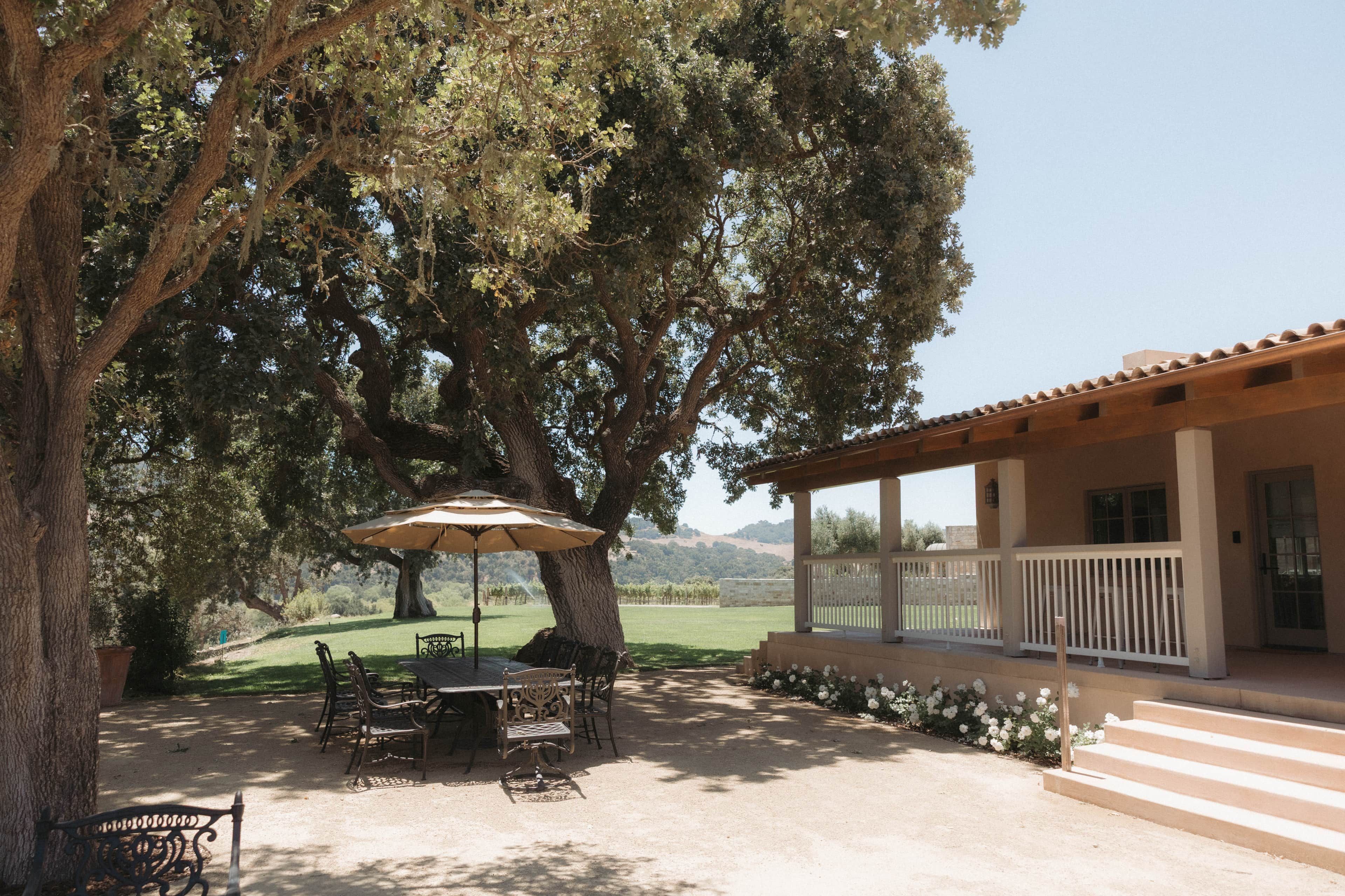 An outdoor patio with a table and chairs, surrounded by a large tree and a building with a balcony.