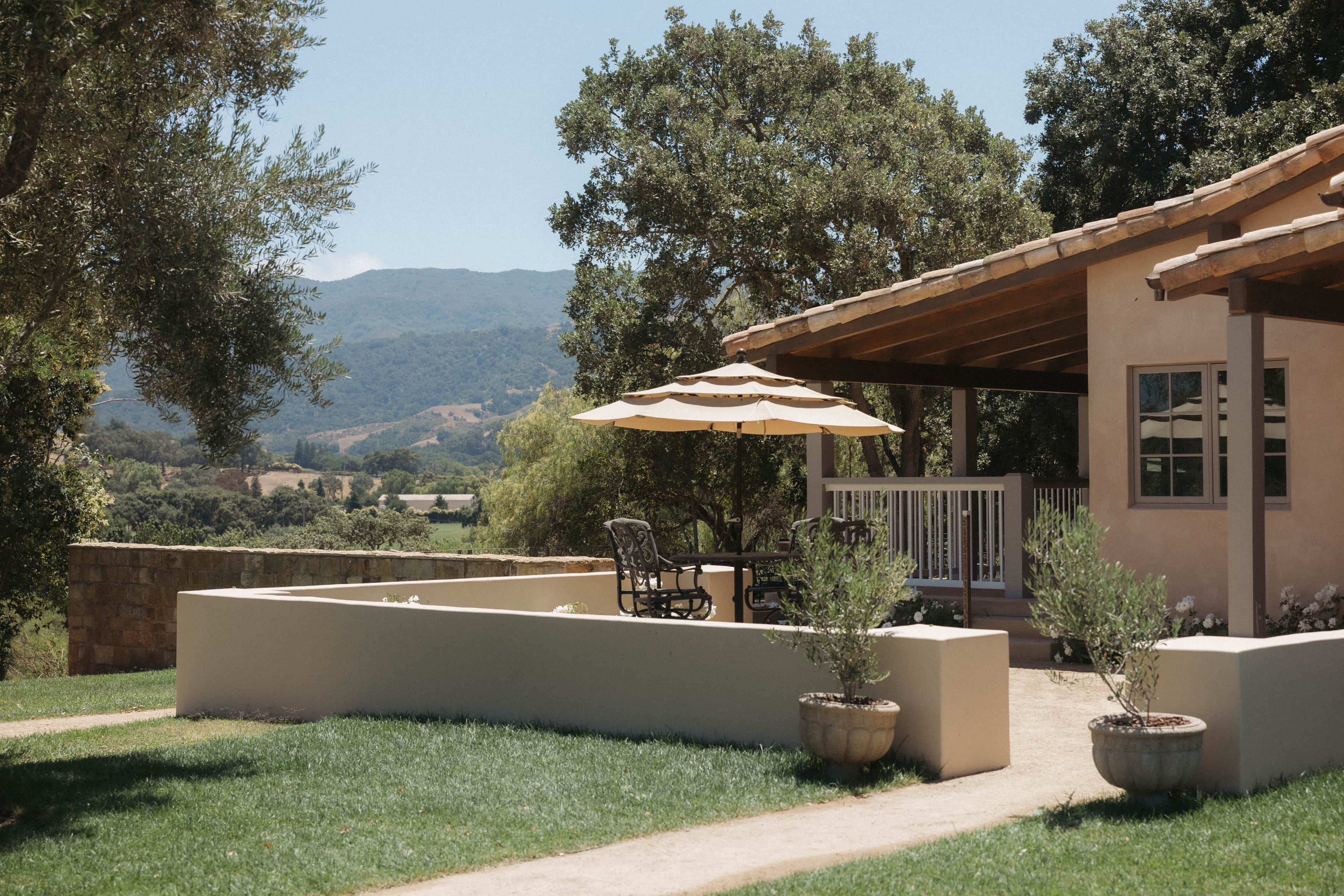 An outdoor patio area with a table and chairs, a pergola with an umbrella, and a view of the surrounding landscape.
