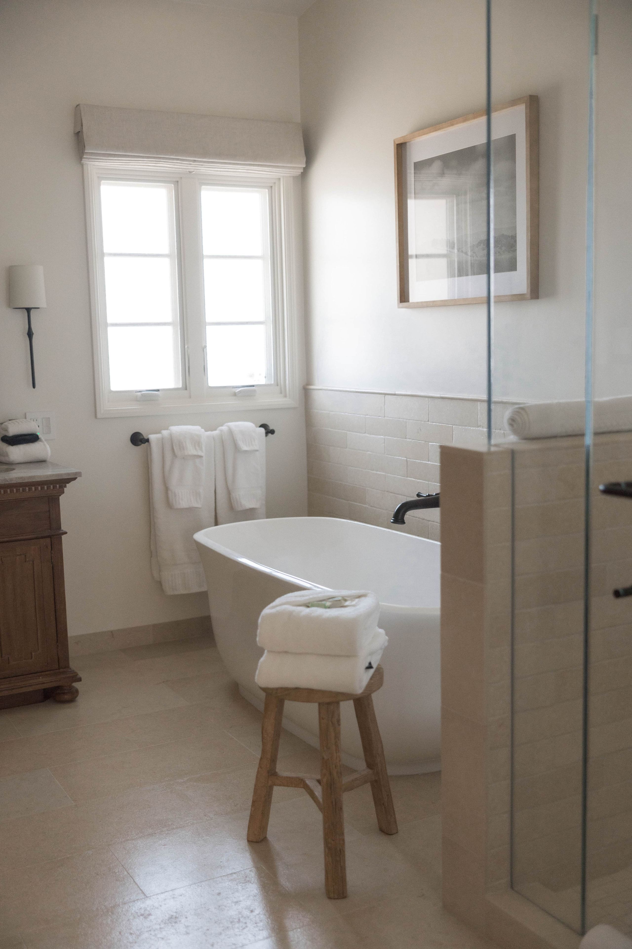 A bathroom with a bathtub, a wooden stool, a window with curtains, and a framed picture on the wall.