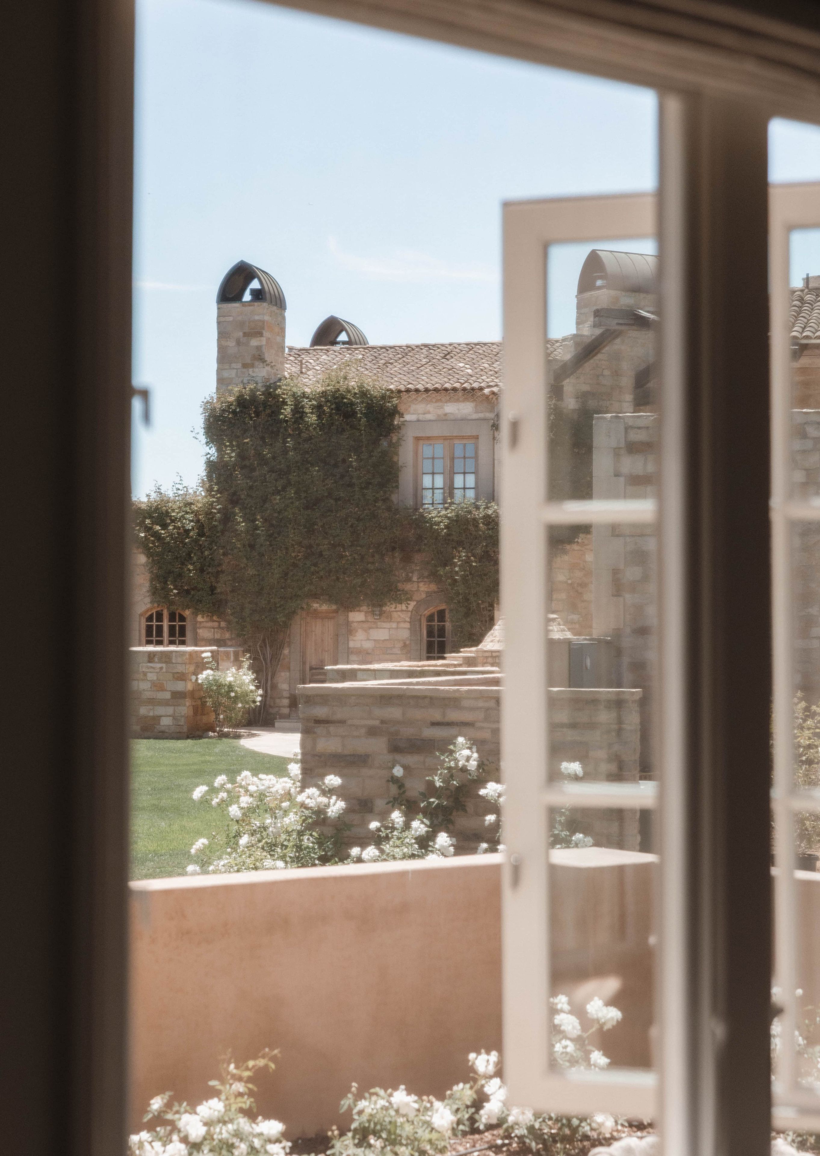 Vista a través de una ventana de un edificio de piedra con techo de tejas, chimenea y un jardín con flores blancas.