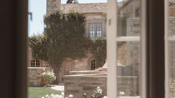 Vista a través de una ventana de un edificio de piedra con techo de tejas, chimenea y un jardín con flores blancas.