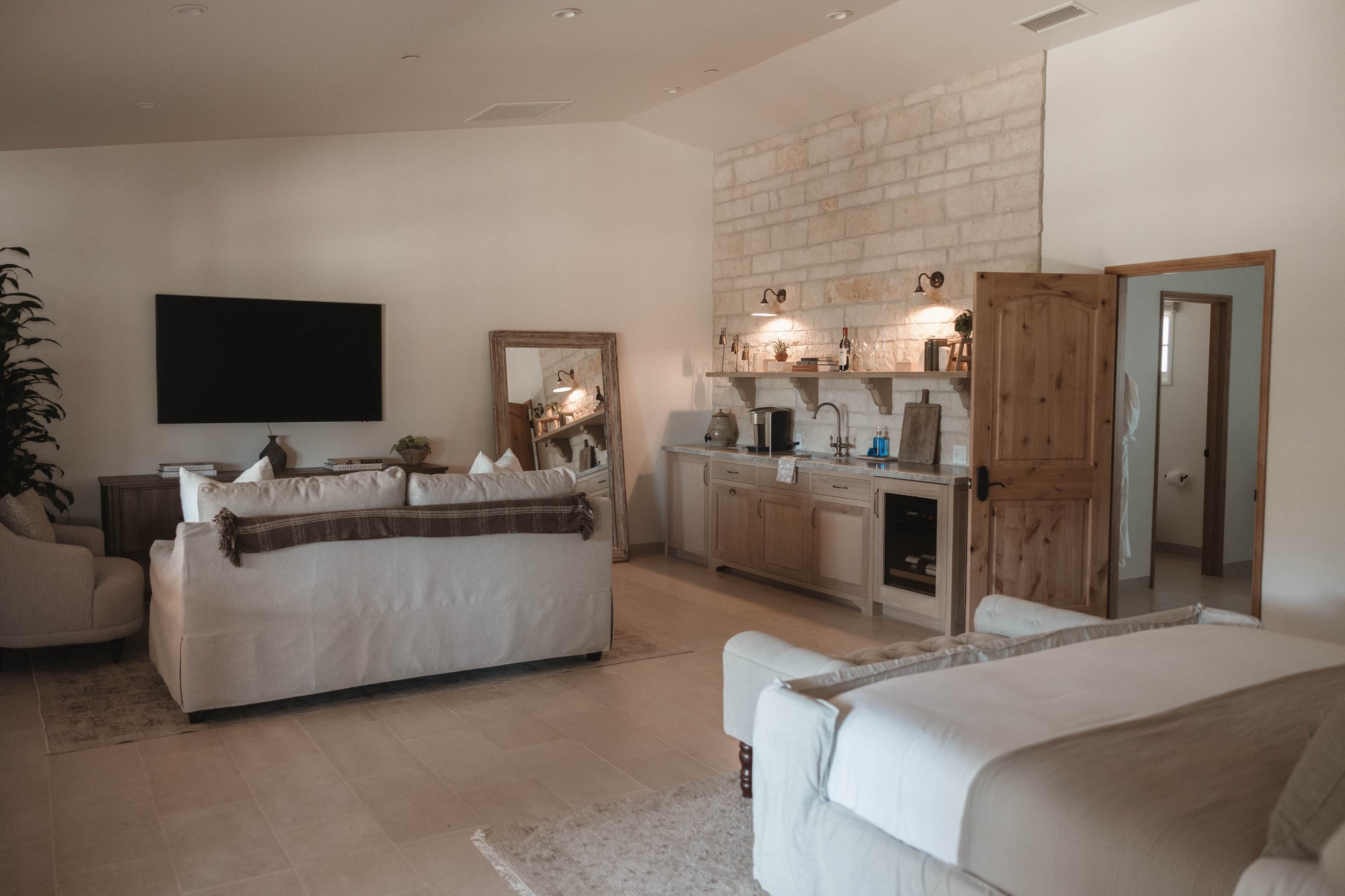Una sala de estar con sofá, mesa de centro, televisor y una zona de cocina con pared de ladrillo y muebles de madera.