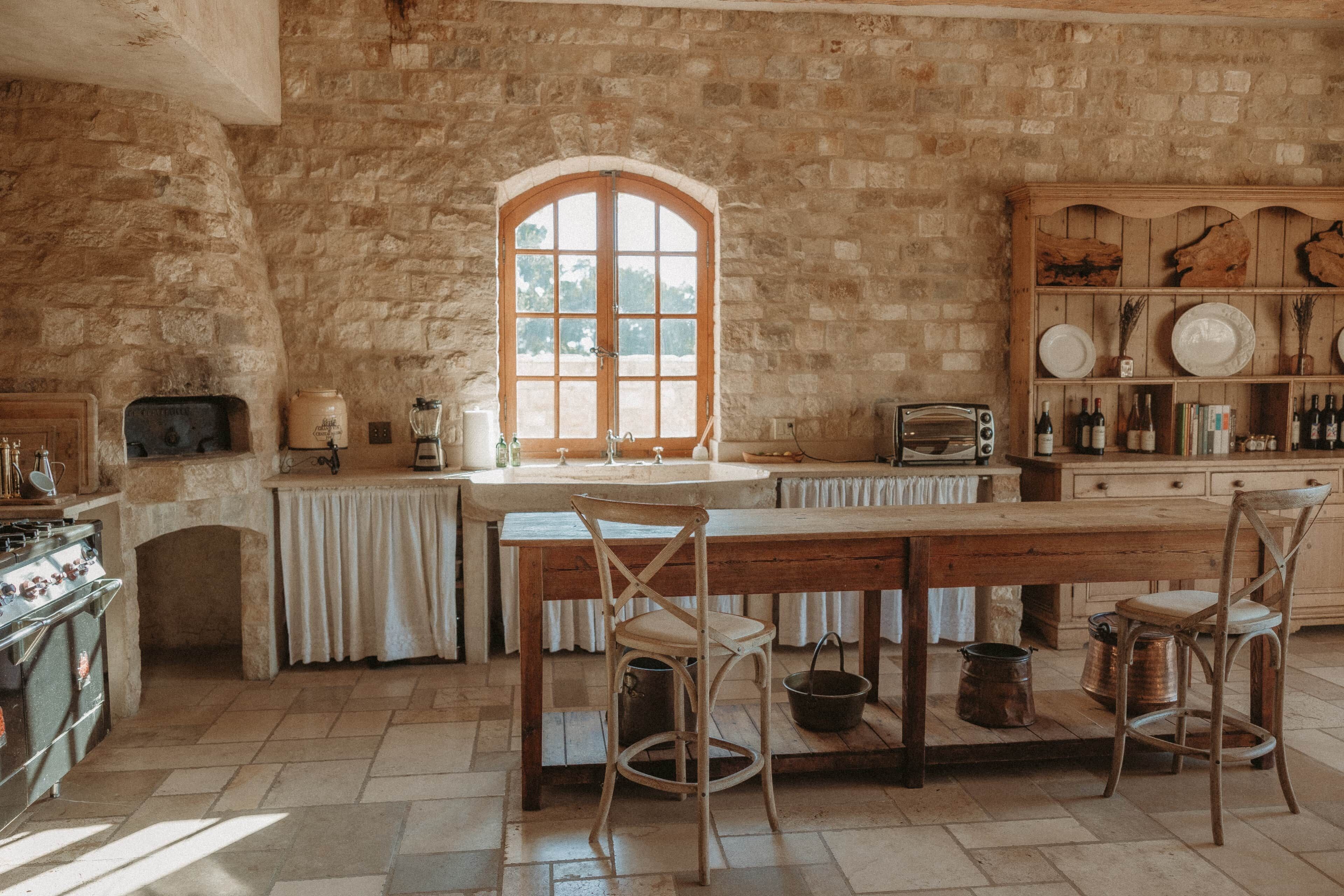 A rustic kitchen with stone walls, a wooden table, and a large arched window.