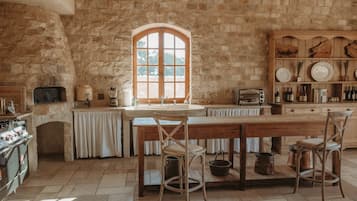A rustic kitchen with stone walls, a wooden table, and a large arched window.