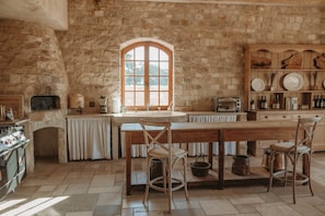 A rustic kitchen with stone walls, a wooden table, and a large arched window.