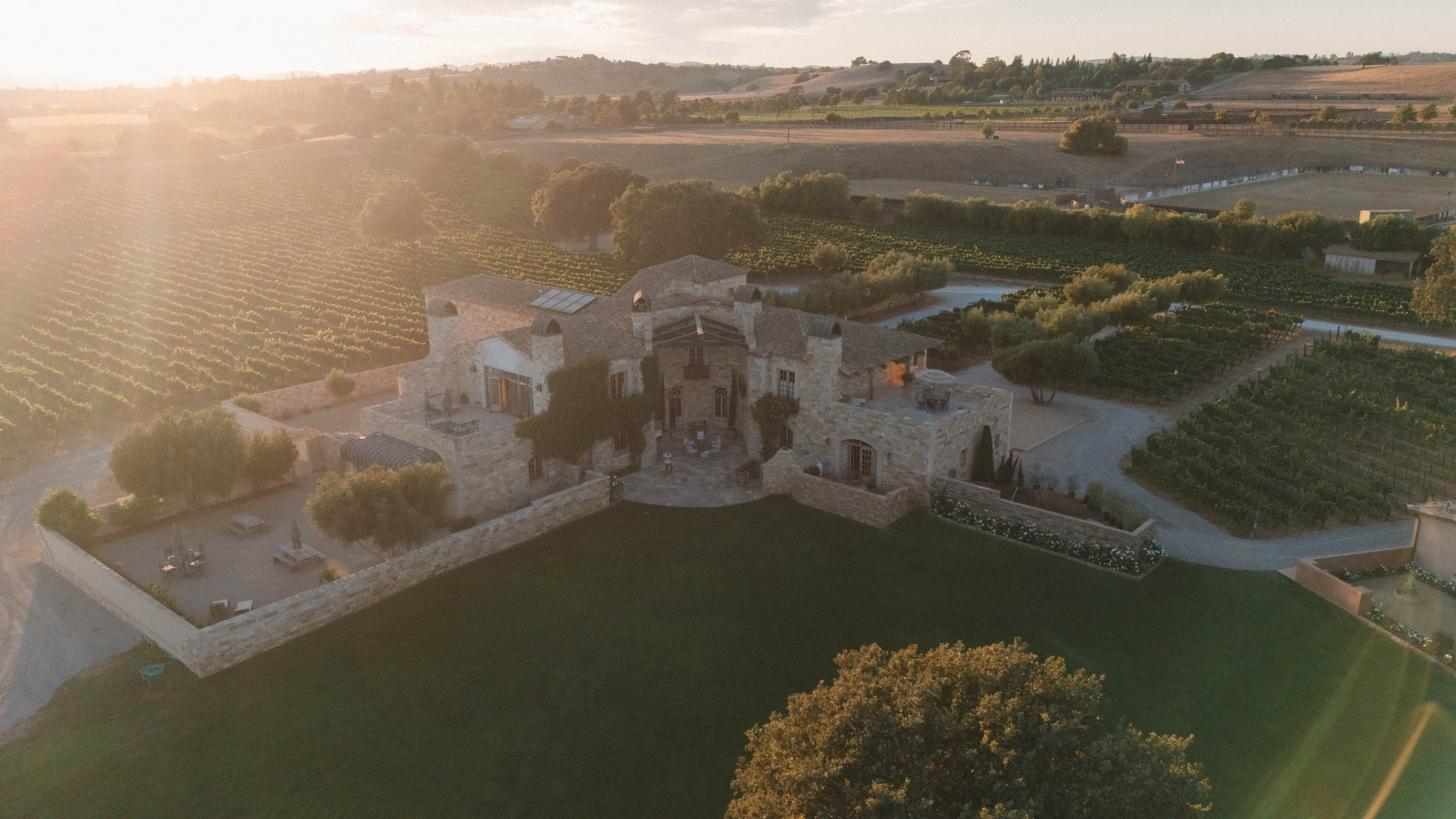 An aerial view of a historic stone building with a courtyard, surrounded by vineyard fields.