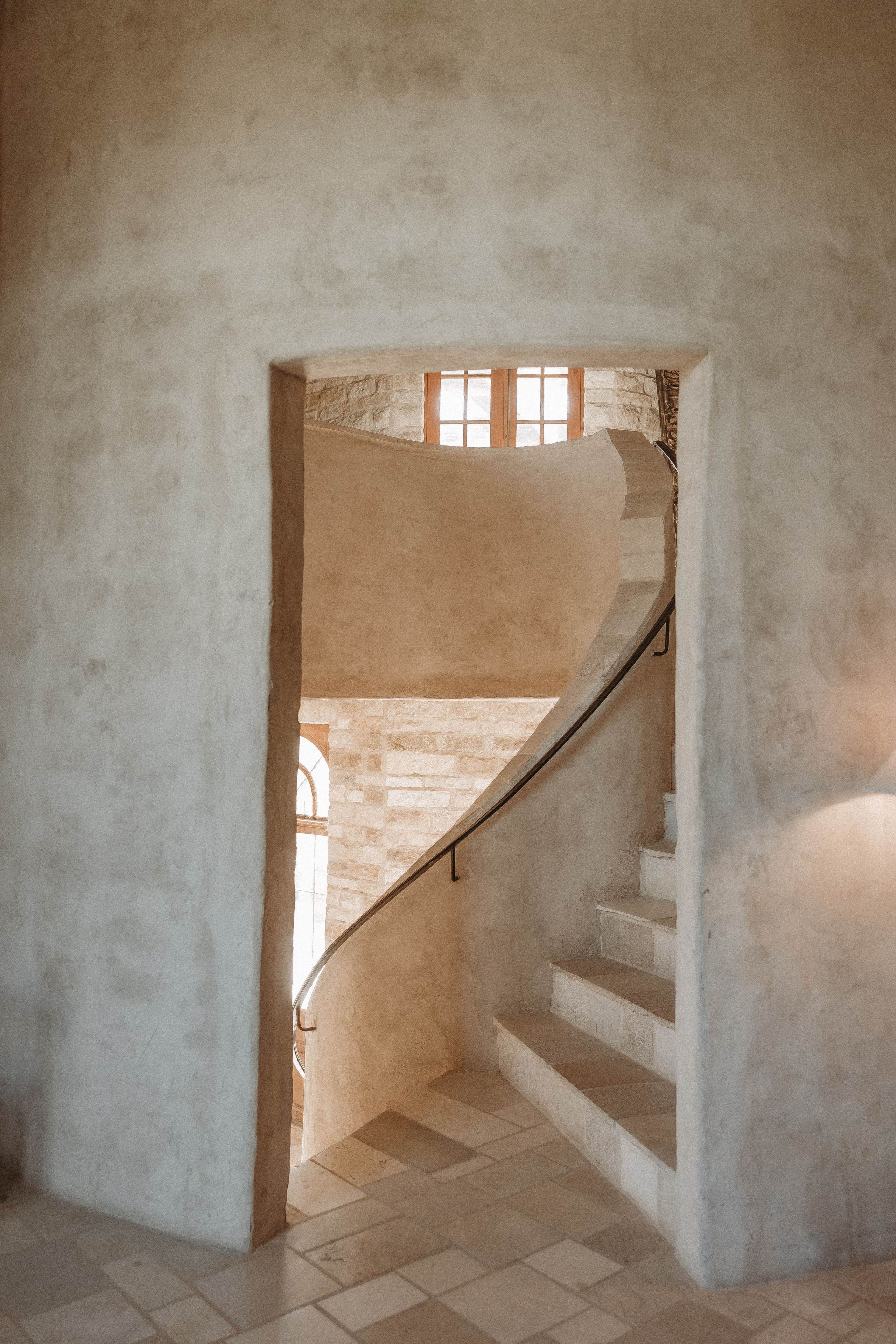 A spiral staircase leading to an upper floor, with a stone wall and a window in the background.
