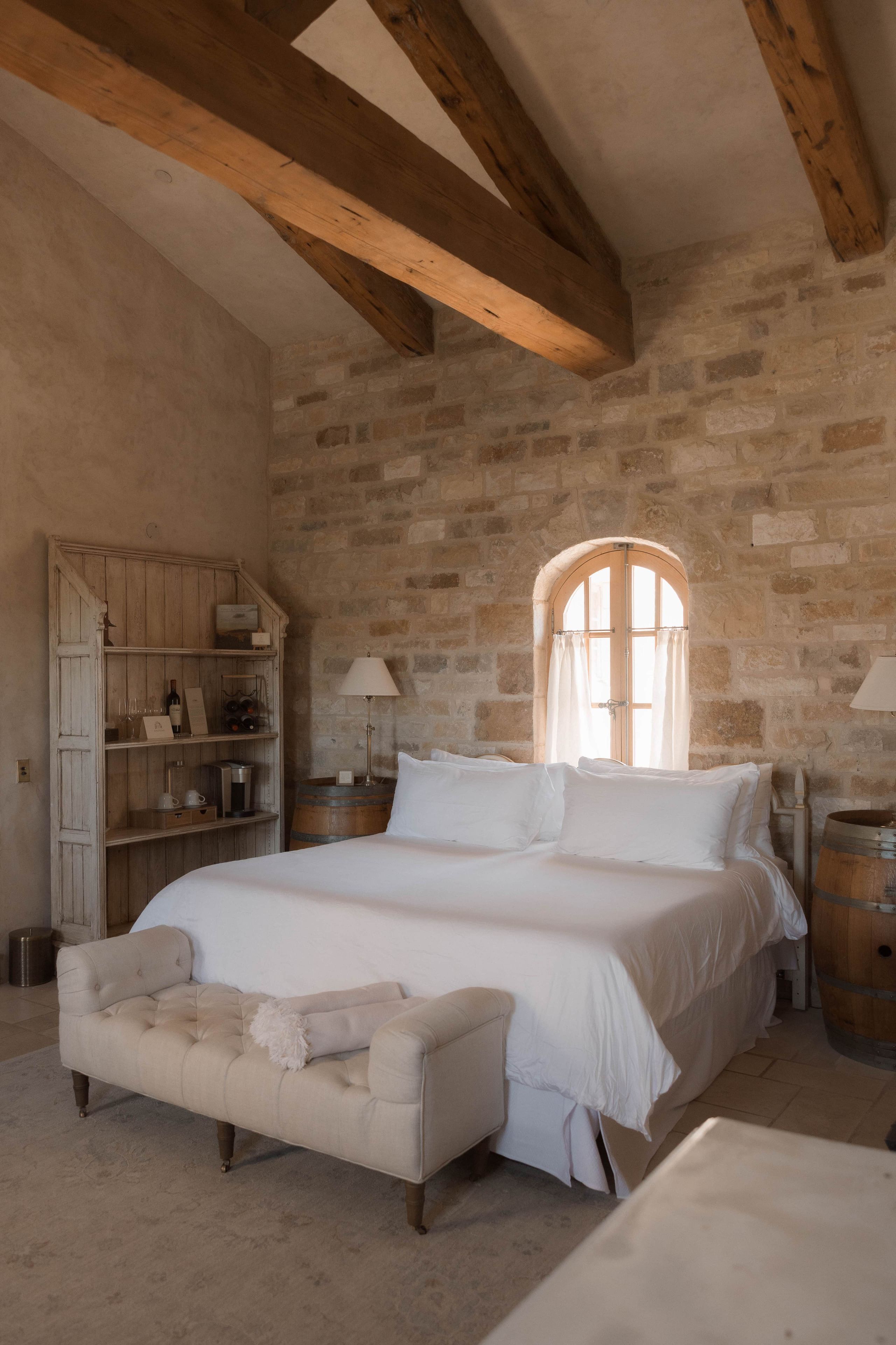 A bedroom with a large bed, wooden beams, stone walls, a wooden cabinet, and a window.