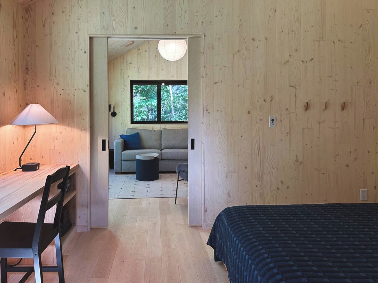 A room with a wooden interior, a bed, a desk with a chair, a sofa, and a window with a view of trees.
