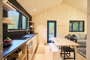 A modern kitchen with a built-in stove, a dining area with a bench, and a view of the outdoors through a door. - Scandi Spruce Cabin in the Woods #2 (Grand Rapids)