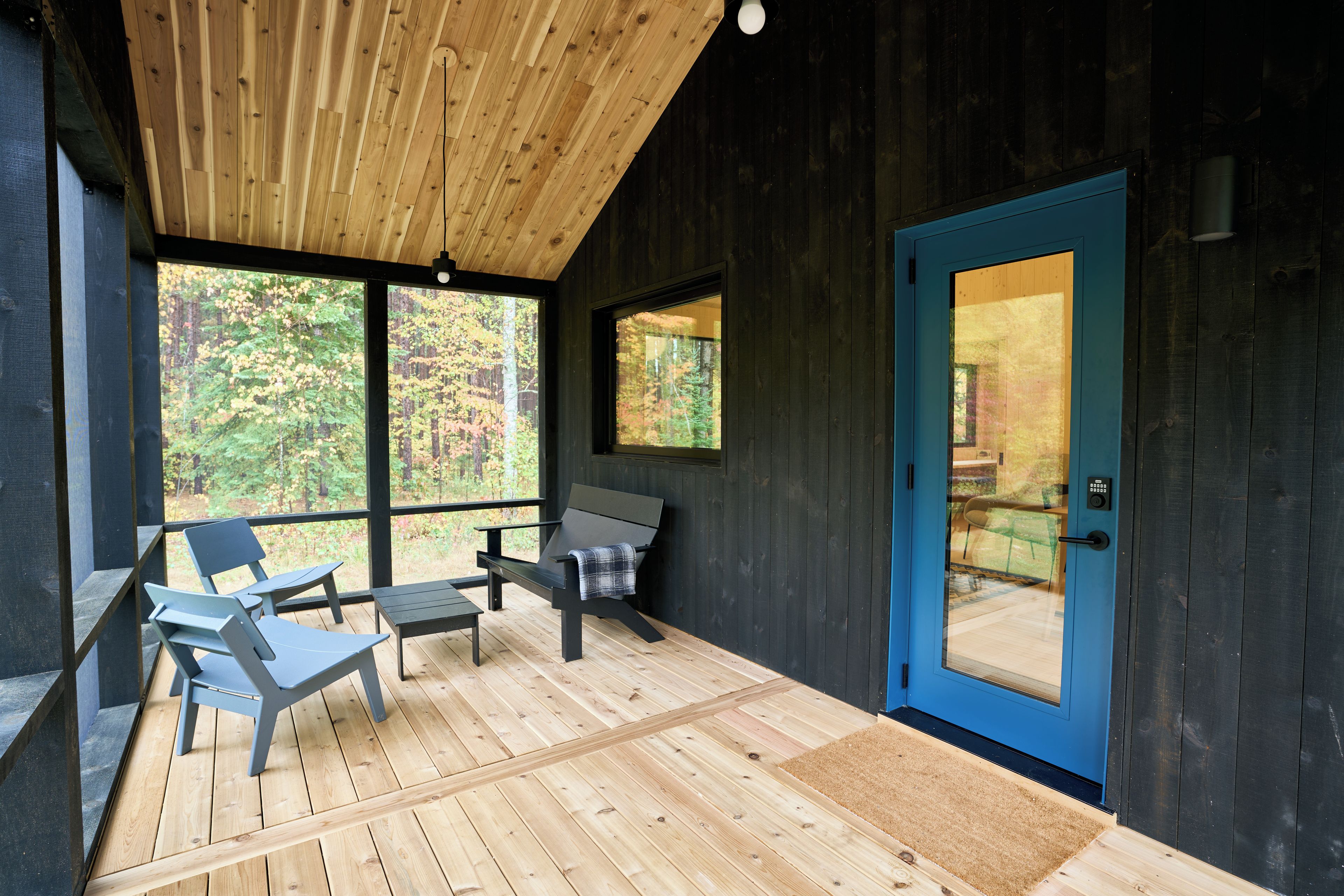 A wooden deck with two chairs and a bench, a blue door, and large windows overlooking a forest.