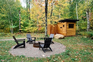 A wooden cabin with a small deck, surrounded by a forest with autumn foliage. There are four Adirondack chairs arranged around a fire pit. - Scandi Spruce Cabin in the Woods #2 (Grand Rapids)