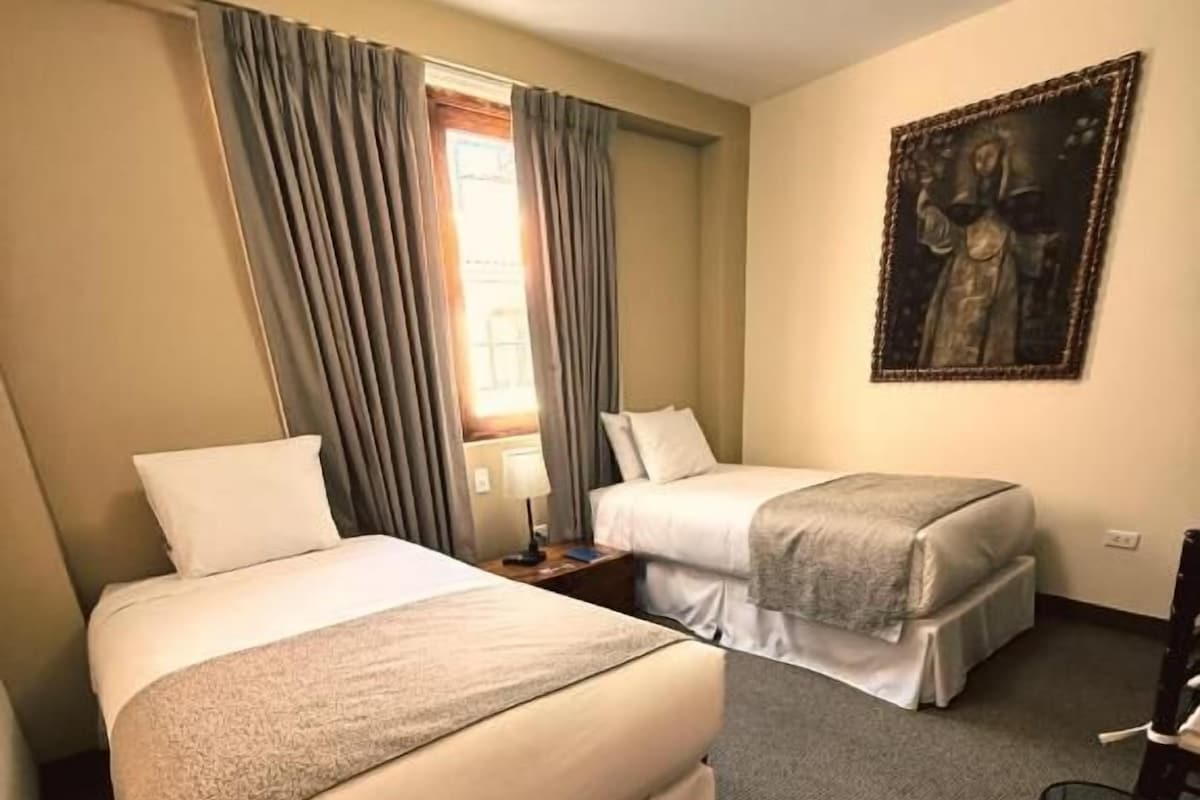 Quarto de hotel com duas camas, janela com cortinas, um quadro na parede e uma mesinha com luminária.
