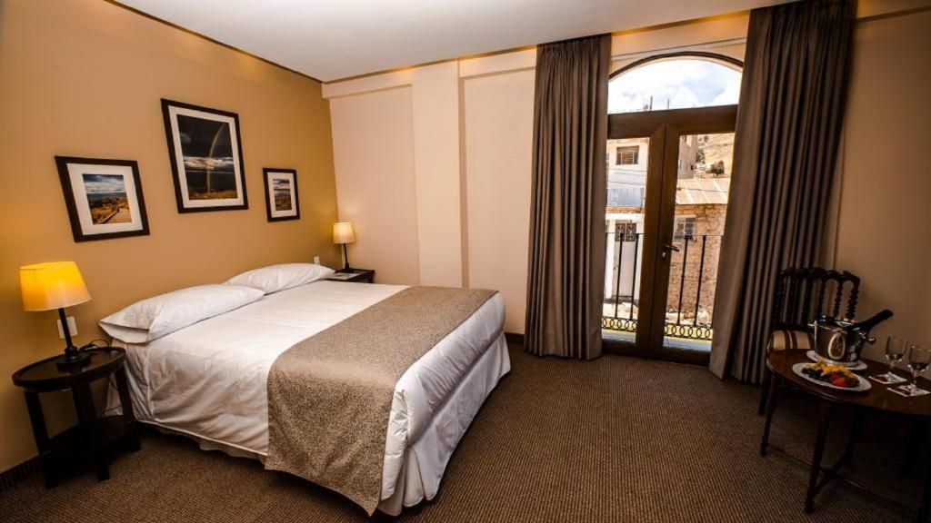 A hotel room with a bed, a nightstand, a lamp, a dining table with a plate and glasses, and a view through a door.