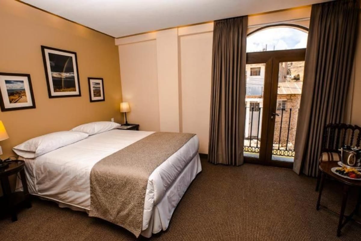 Quarto de hotel com cama, mesa de cabeceira, abajur, mesa de jantar com prato e copos, e vista através de uma porta.