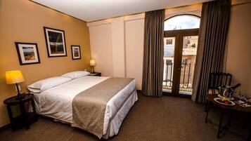 A hotel room with a bed, a nightstand, a lamp, a dining table with a plate and glasses, and a view through a door.