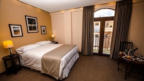 Quarto de hotel com cama, mesa de cabeceira, abajur, mesa de jantar com prato e copos, e vista através de uma porta.