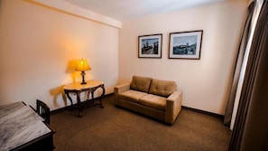 Quarto de hotel com sofá, uma pequena mesa com abajur e duas quadros na parede.