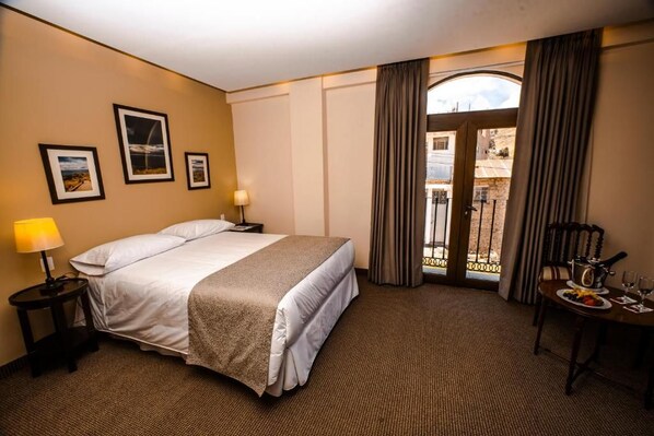 A hotel room with a bed, a nightstand, a table with a plate of food and a wine glass, and a balcony with curtains.