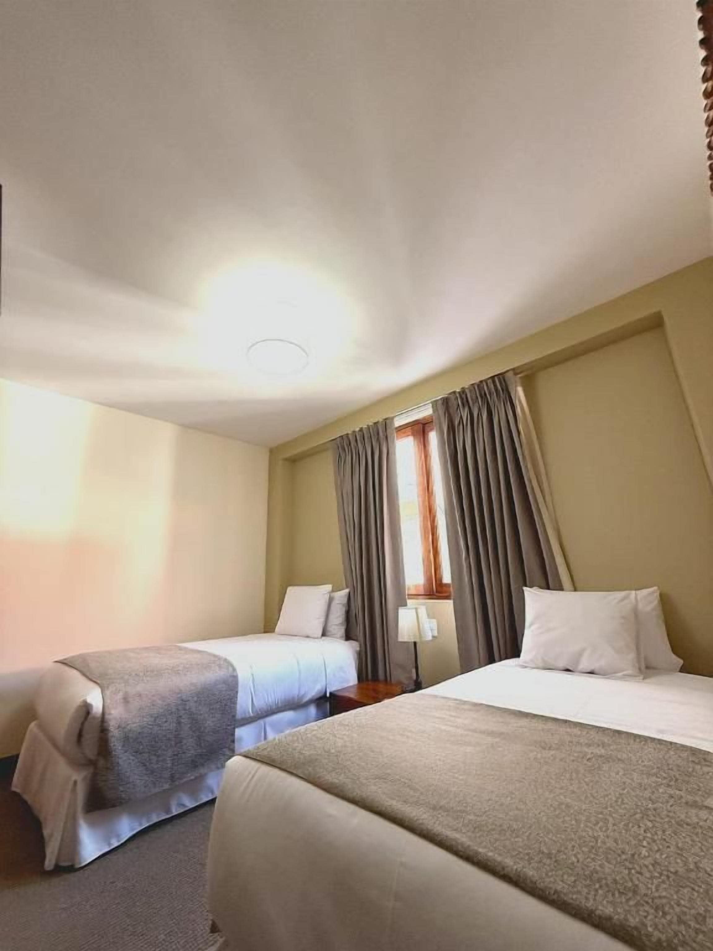 Ein Hotelzimmer mit zwei Betten, einem Fenster mit Vorhängen, einem Nachttisch und einer Deckenleuchte.