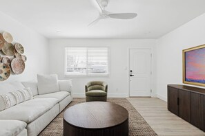Un salon moderne avec un canapé blanc, une table basse ronde, un fauteuil vert, un ventilateur de plafond, une fenêtre avec des stores, une porte blanche et un tableau encadré accroché au mur.