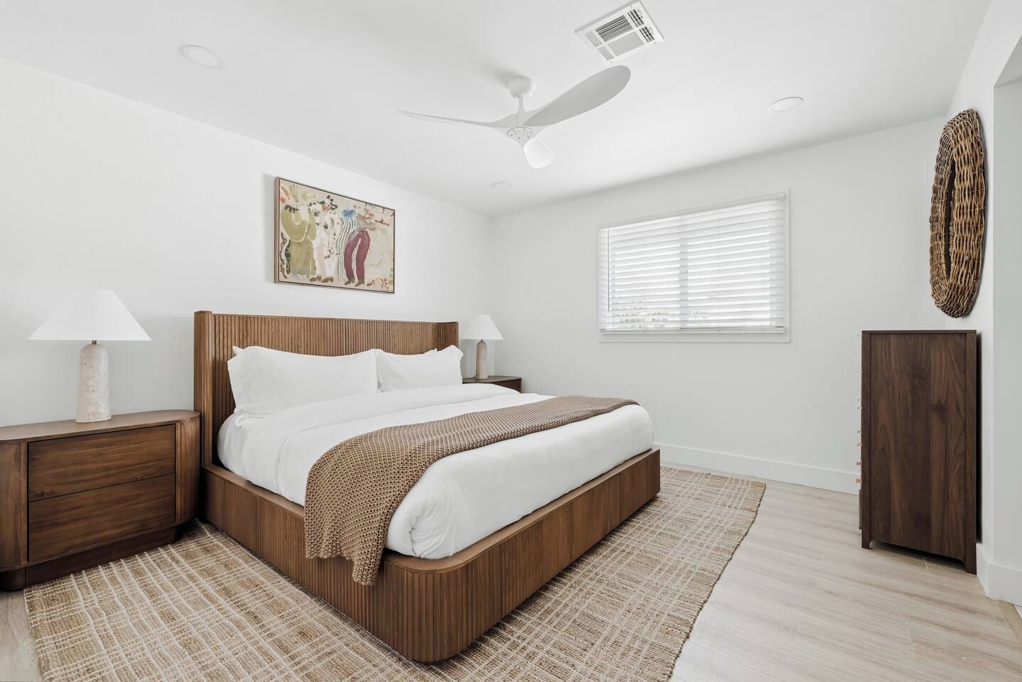 A bedroom with a bed, nightstands, a ceiling fan, a window with blinds, and a wall-mounted artwork.