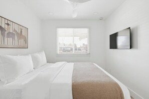 Une chambre à coucher avec un lit, une fenêtre avec des stores, un ventilateur de plafond et un téléviseur fixé au mur.