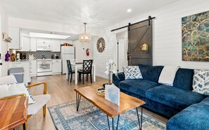 A modern living room with a blue sofa, wooden coffee table, and a dining area with chairs. - 5 Stars~WALK TO BEACH from Serendipity Suites! 2-of-2  2-bed/2-ba Cottage Suites (Holmes Beach)