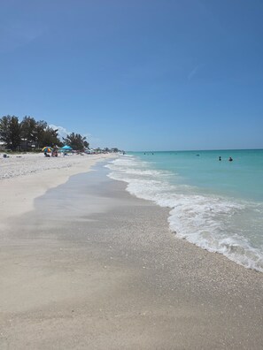 Ein Strand mit blauem Himmel, ruhigem Wasser und einigen badenden Personen.