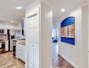 A kitchen with white cabinetry, a refrigerator, and a microwave. There is a door leading to a hallway with a blue accent wall and framed artwork. - Sea La Vie- Luxury Ocean View Condo- 1 to 6 month stay (Seaview)