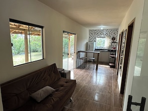 A living room with a brown sofa, a dining area with a table and chairs, and a kitchen with a refrigerator. - House for 6 People - Recanto dos Eucaliptos - Great location - Pet Friendly (Imbituba)