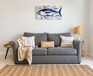 A grey sofa with a blanket and a pillow with text, a small wooden side table with a plant, and a lamp on a side table. - Marathon Key Getaway (Marathon)