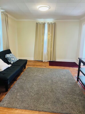 A room with a black sofa, a grey carpet, wooden flooring, beige curtains, and a ceiling light. - Peaceful 2-bedroom house in charming Rutland! Have fun with family & friends (Rutland)