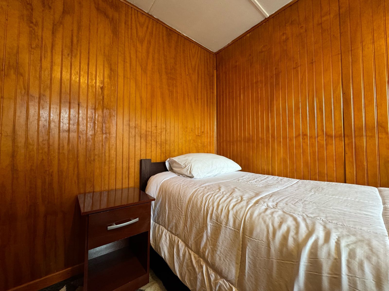 Un dormitorio con pared de paneles de madera, una cama individual con colcha blanca y una mesita de noche de madera con cajón.
