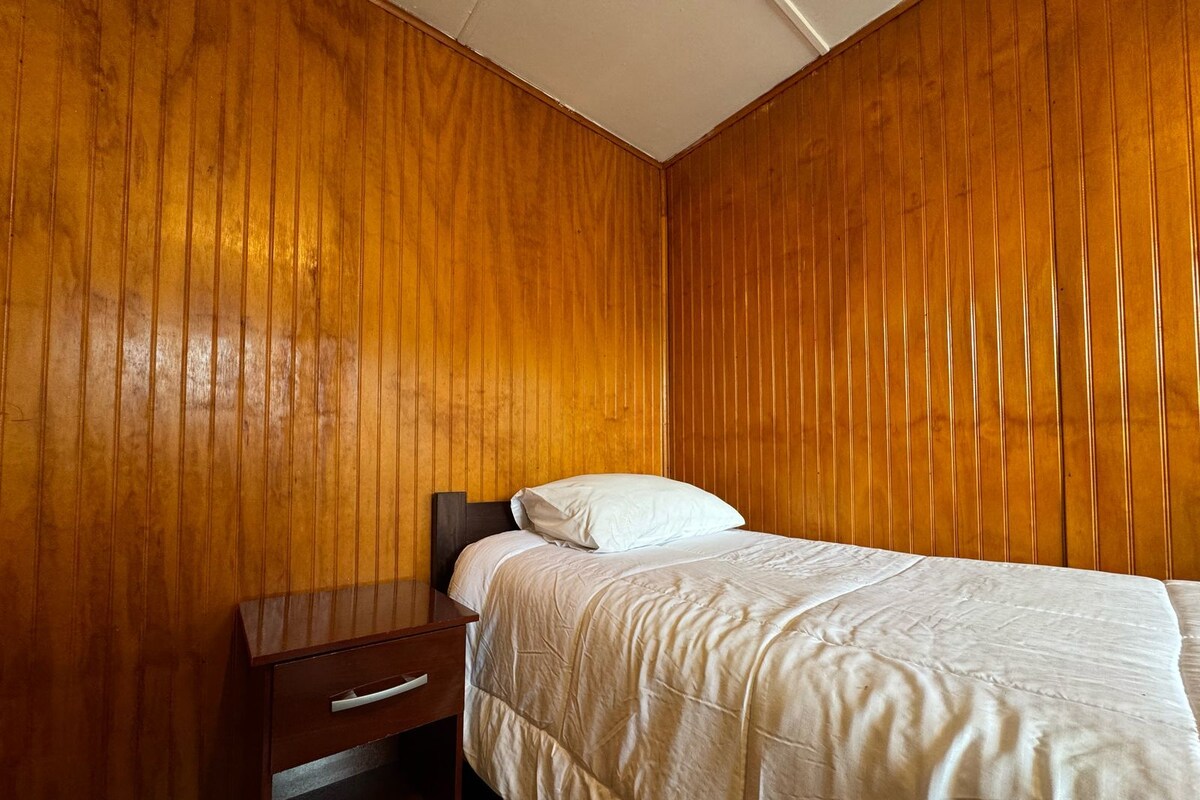 Een slaapkamer met een houten lambrisering, een enkel bed met een witte spreideken, en een nachtkastje van hout met een lade.