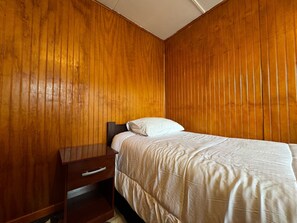 Een slaapkamer met een houten lambrisering, een enkel bed met een witte spreideken, en een nachtkastje van hout met een lade.