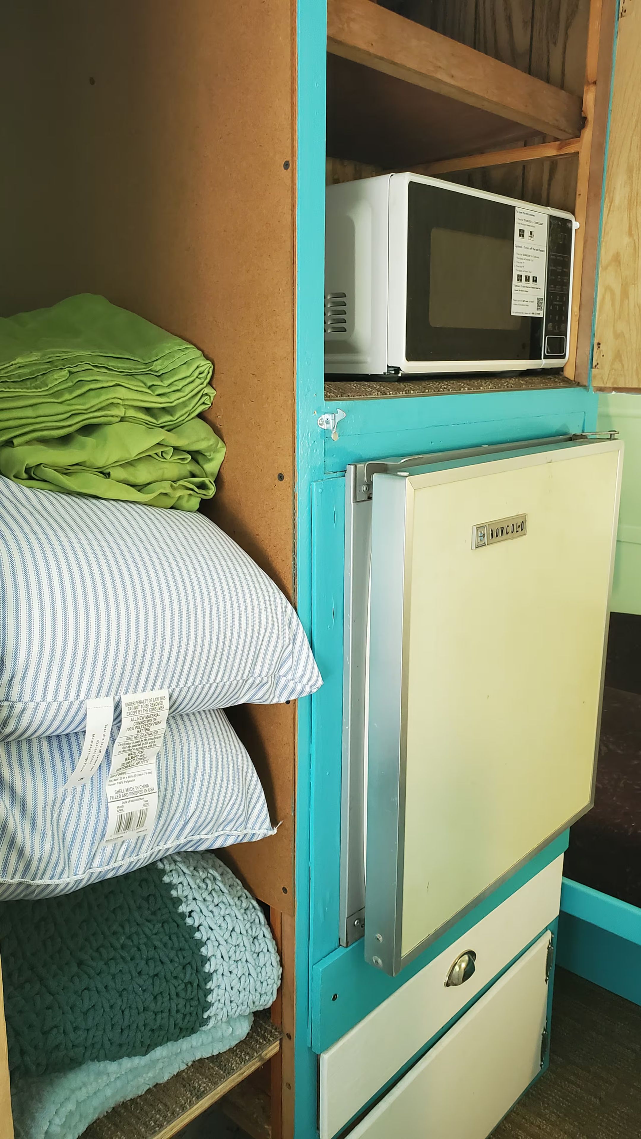 Una kitchenette compacta con microondas, refrigerador y un estante con mantas dobladas.