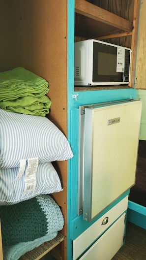 A compact kitchenette with a microwave, a refrigerator, and a shelf with folded blankets. - The -DaLtOn- (Naches)