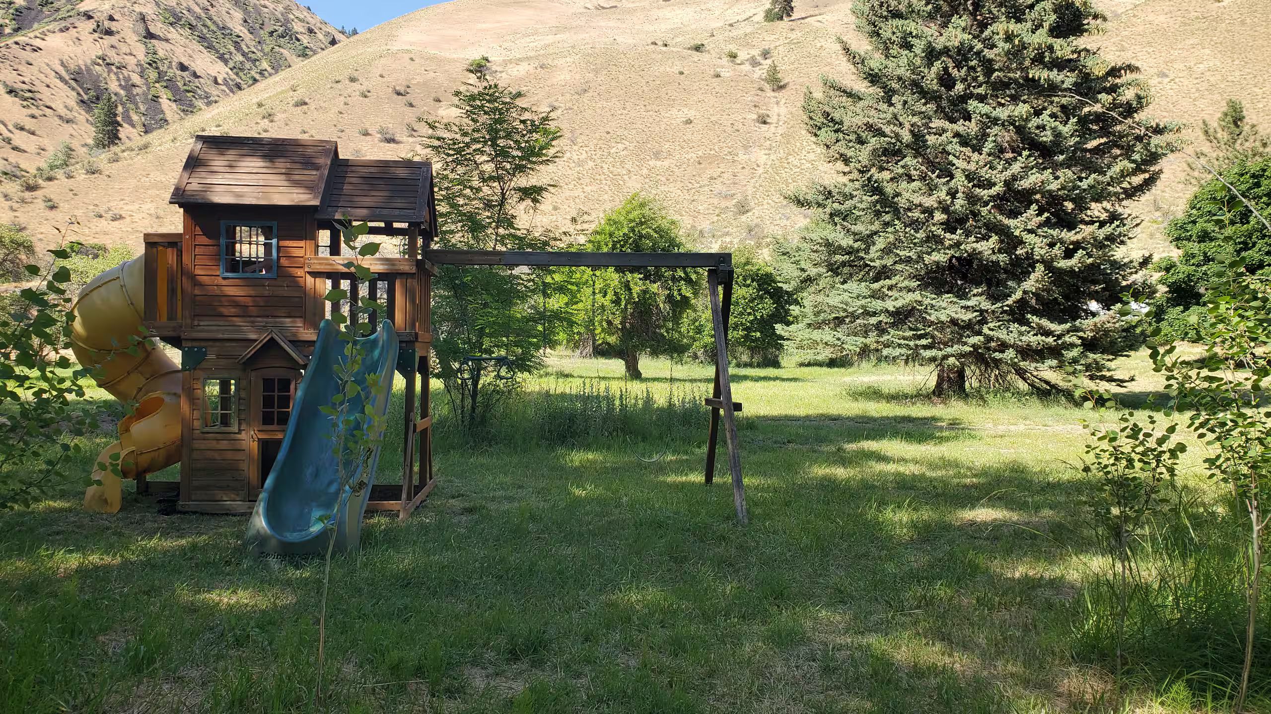 A wooden playhouse with a slide in a grassy field with trees and mountains in the background.