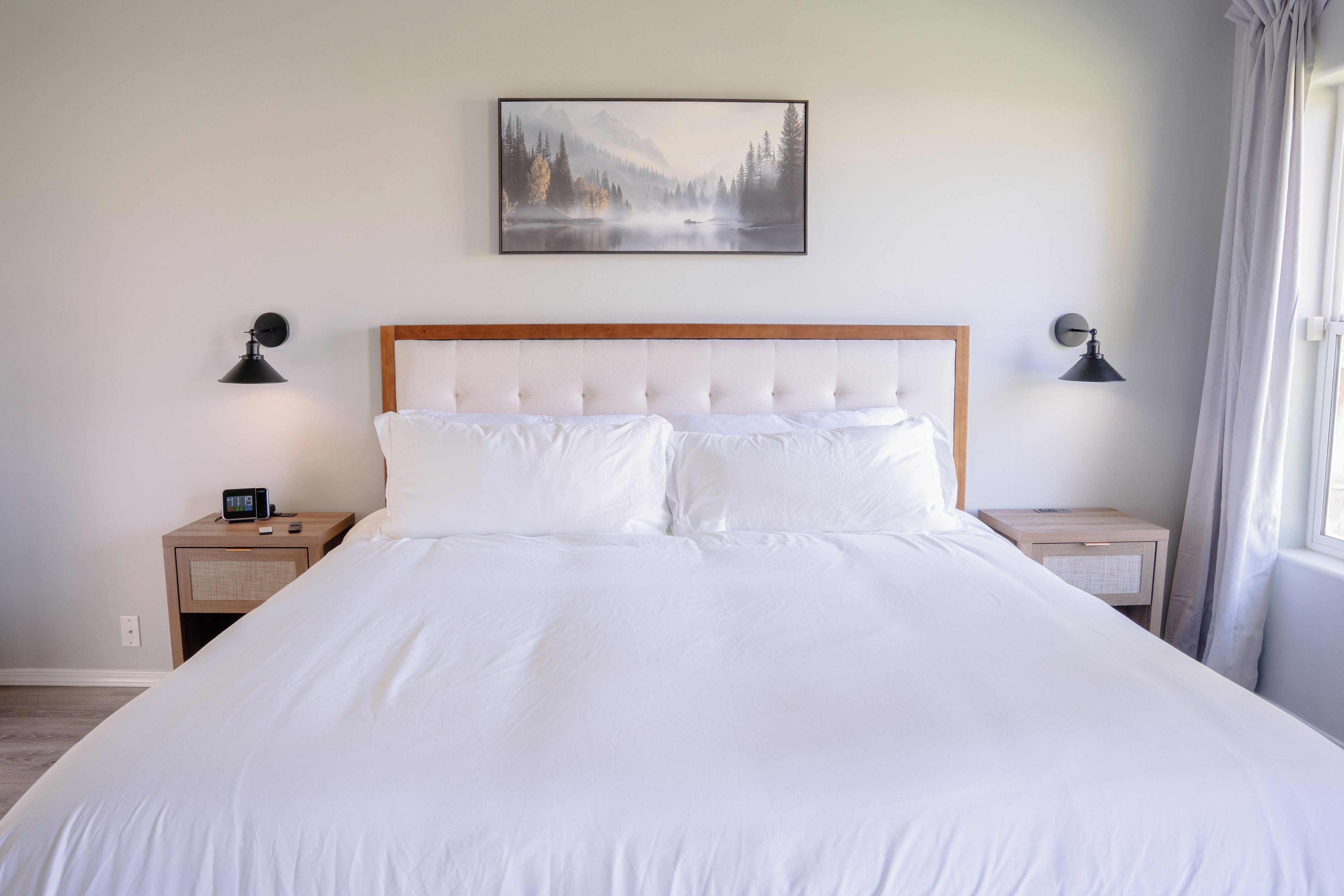 Una cama bien hecha con cabecera de madera, dos mesitas de noche con lámparas, un cuadro colgado en la pared y una ventana con cortinas.