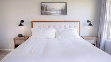 Una cama bien hecha con cabecera de madera, dos mesitas de noche con lámparas, un cuadro colgado en la pared y una ventana con cortinas.