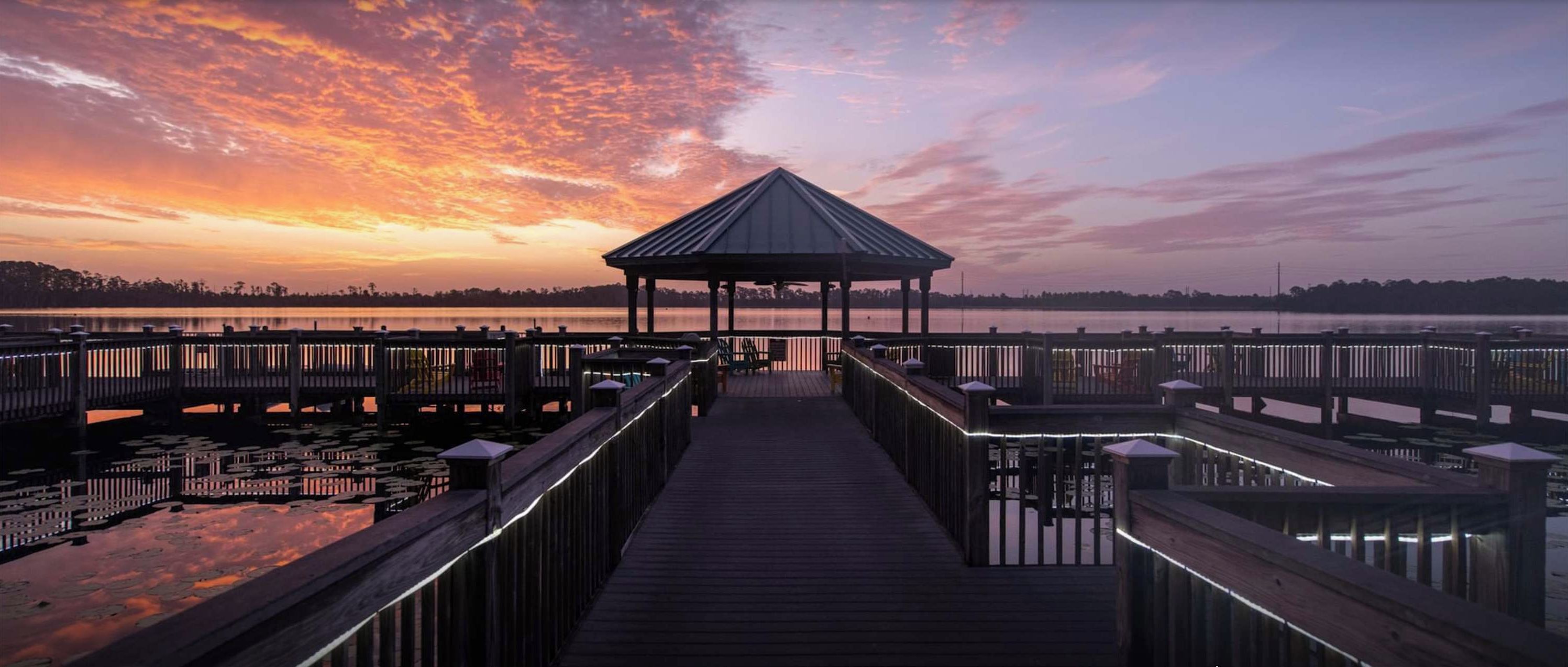 A wooden pier leading to a gazebo by a lake at sunset.