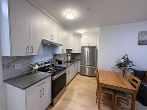 A modern kitchen with grey cabinets, a stainless steel refrigerator, and a wooden dining table. - Spacious 2BR Suite| Walk to White Rock Beach (White Rock)