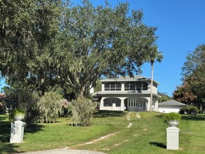 Une maison à deux étages avec une grande véranda à l’avant et un arbre dans le jardin.