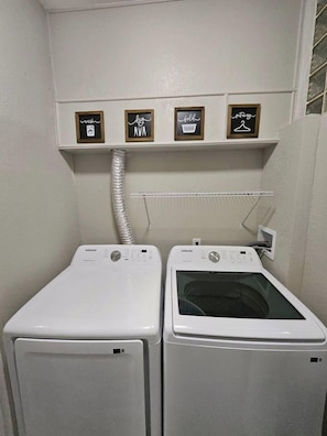 A laundry area with two white washing machines and a shelf above them with four framed signs. - Beautiful Historical 6 Bdrm Lakefront Pool Home (Auburndale)