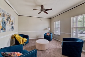A room with a ceiling fan, two blue armchairs, a round ottoman, and a window with blinds. - The Westfield Retreat | 7 Bedrooms • 30+ Day Stays (Westfield)