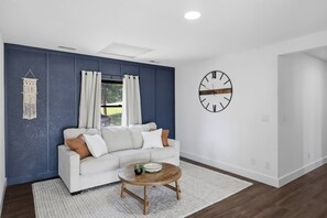 A living room with a white sofa, a round wooden coffee table, a macrame wall hanging, and a large wall clock. - Booje Bay : Luxury, Lakefront, Hot Tub! (Sunrise Beach)