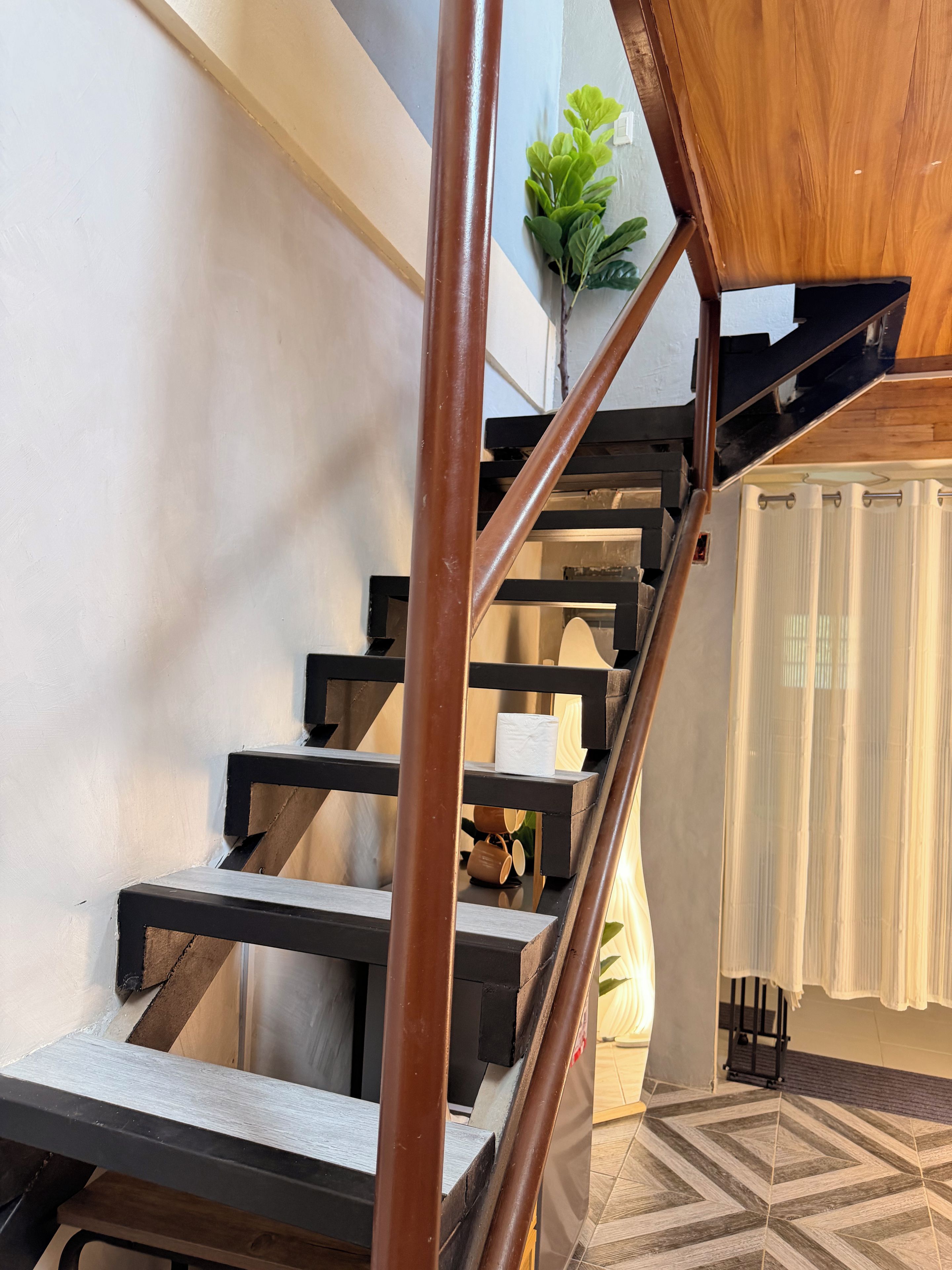 A staircase with wooden steps and metal railings, leading to a room with a patterned floor and a plant in the corner.