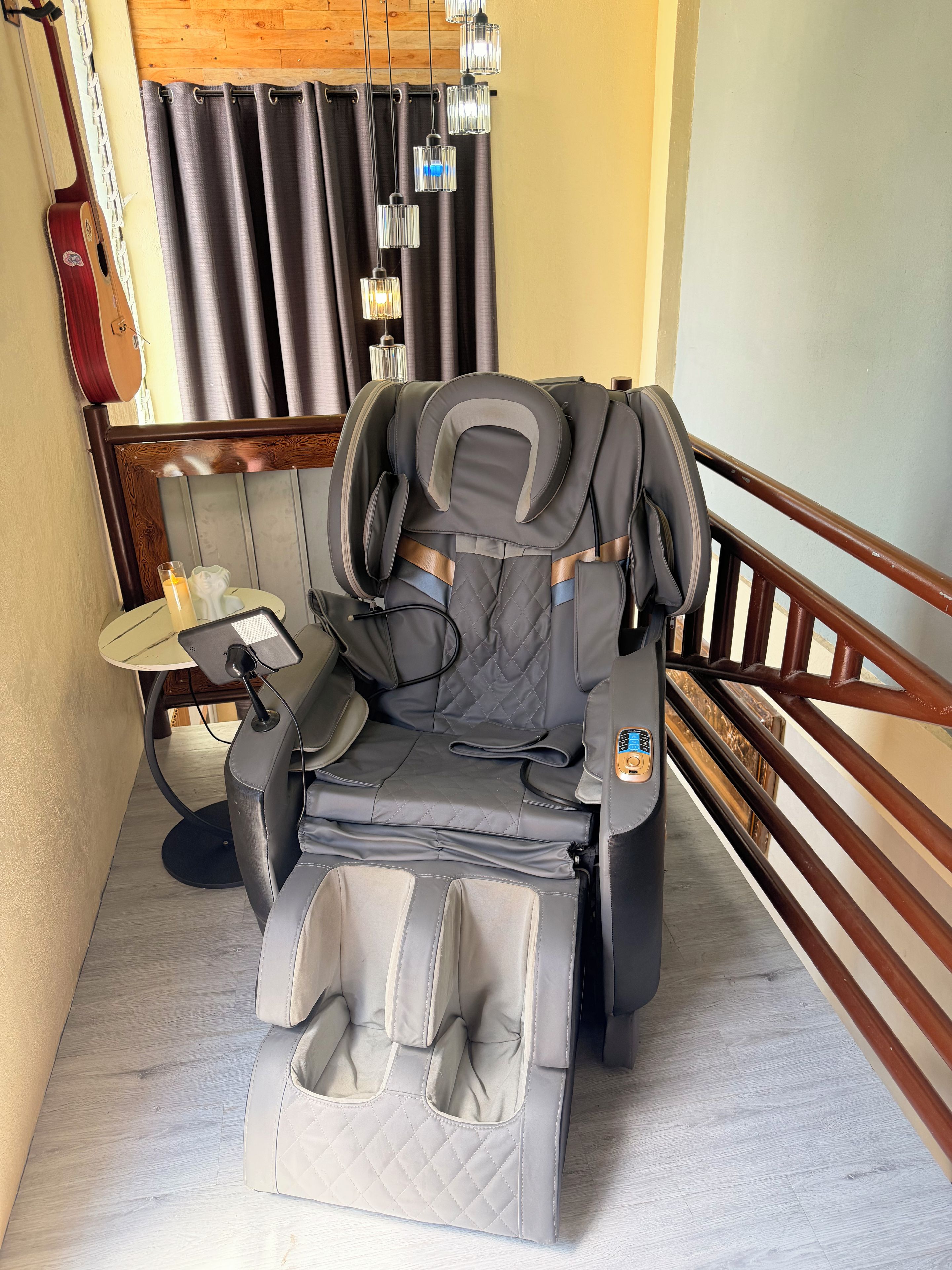 A room with a massage chair, a table with a lamp, and a window with curtains.