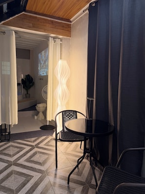 Una stanza con una sedia in metallo nero, un piccolo tavolo rotondo, un lampadario da terra bianco e vista su un bagno attraverso una porta aperta.